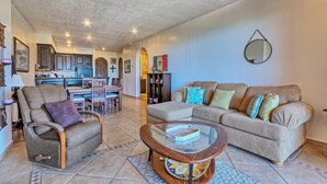 A living room with a sofa, armchair, and coffee table, a kitchen in the background, and a dining area.