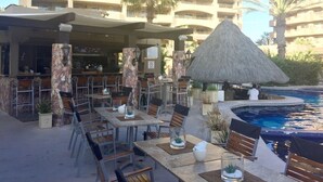 An outdoor dining area with tables and chairs, a thatched-roof structure, and a pool in the background.