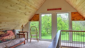 Ein gemütliches Dachzimmer mit Holzbalkendecke, einem Sofa, einem Sessel und einem kleinen Tisch. Große Fenster bieten Blick auf eine grüne Landschaft.