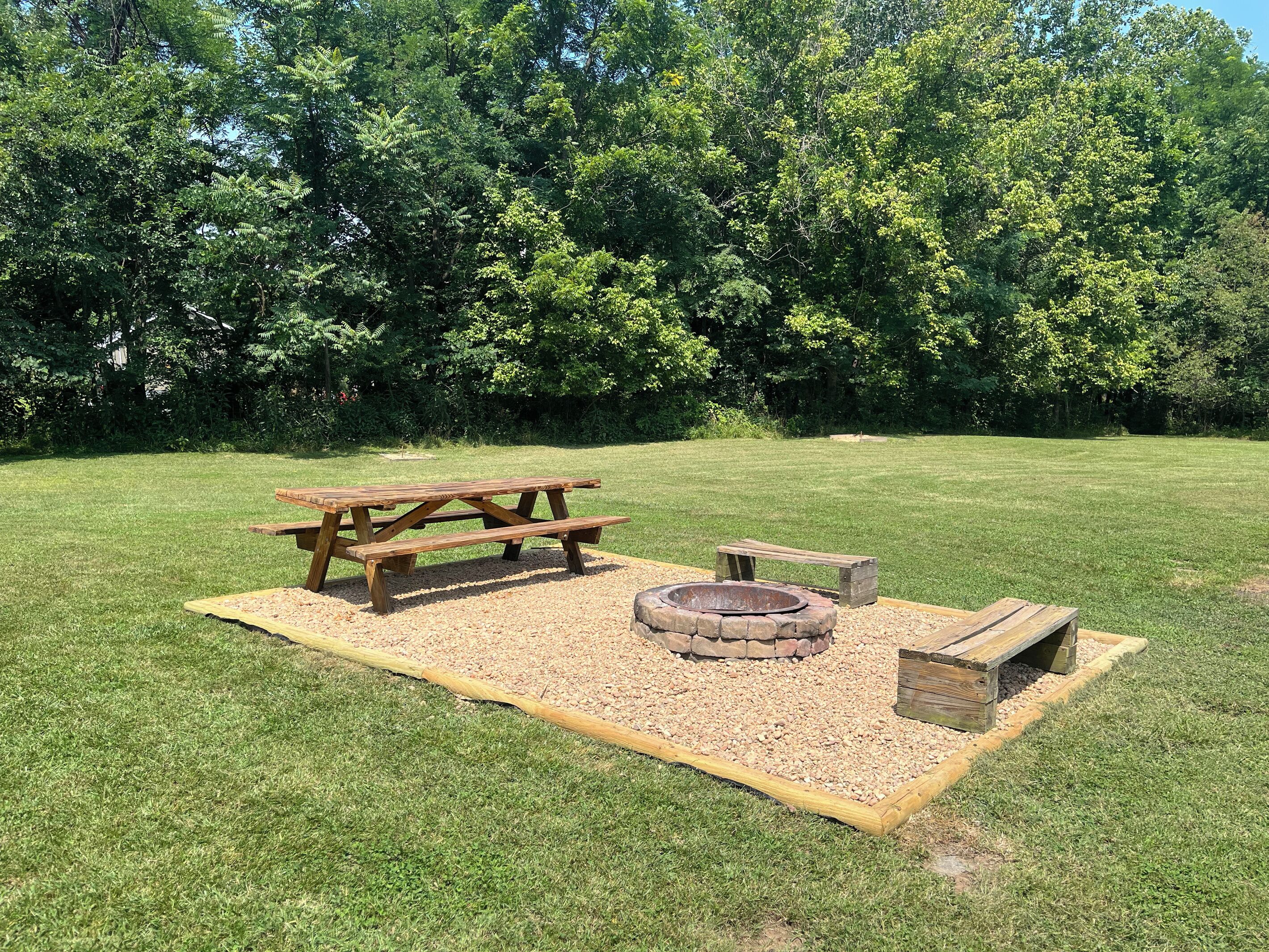 A picnic setup with a wooden table and benches, a fire pit, and a small wooden structure.