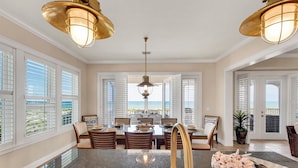A spacious dining area with a view of the ocean, featuring a large table set for dining, white shutters on the windows, and a ceiling with two large pendant lights. - Dolphin Watch | Beachfront on private Beach (Cape Haze)