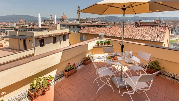 Una terraza en la azotea con una mesa, sillas y una sombrilla. Se ven plantas en macetas y una vista panorámica de la ciudad al fondo.