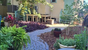 Een tuin met een pad van stenen, diverse planten in potten, een houten terras en een overdekte barbecuehoek.