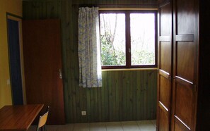 A room with a wooden floor, a window with a curtain, a wooden door, and a table with a chair.