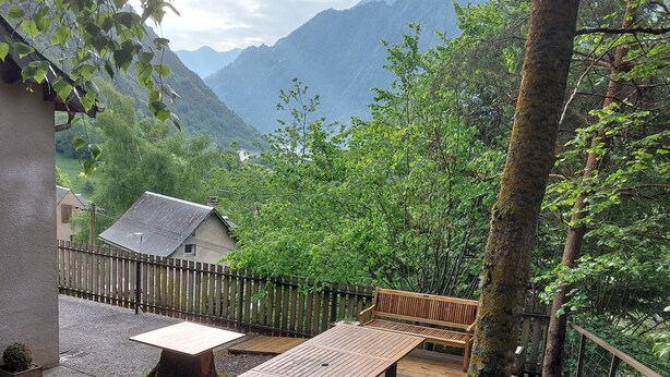 Een houten terras met een tafel en banken, omgeven door bomen en uitzicht op een berg.