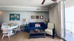 A living room with a ceiling fan, a blue sofa, a coffee table, and a dining area with a white table and chairs. - Beachfront w/ Gulf View | 4th Floor | Elegantly Cleaned | Free WiFi |  (Panama City Beach)