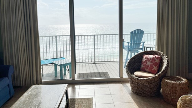 A balcony with a view of the ocean, a wicker chair, a blue chair, and a small table. - Beachfront w/ Gulf View | 4th Floor | Elegantly Cleaned | Free WiFi |  (Panama City Beach)