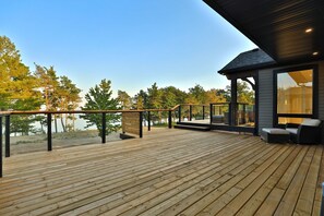 A wooden deck with a railing, outdoor seating, and a view of trees and water. - Waterfront Lake Erie near Niagara Wine Region (Dunnville)