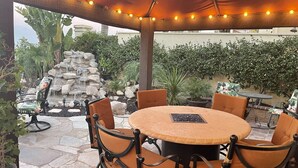 Une terrasse extérieure avec une table ronde et des chaises, une fontaine en pierre et une pergola illuminée de guirlandes lumineuses.
