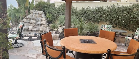 An outdoor patio with a round table and chairs, a stone fountain, and a pergola with string lights.