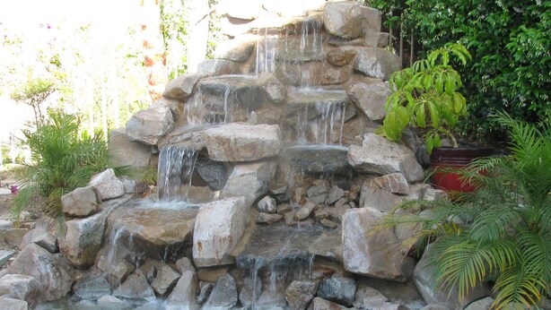 Une cascade artificielle en roche, aménagée dans un jardin où l’on trouve diverses plantes, avec un petit bassin à sa base.