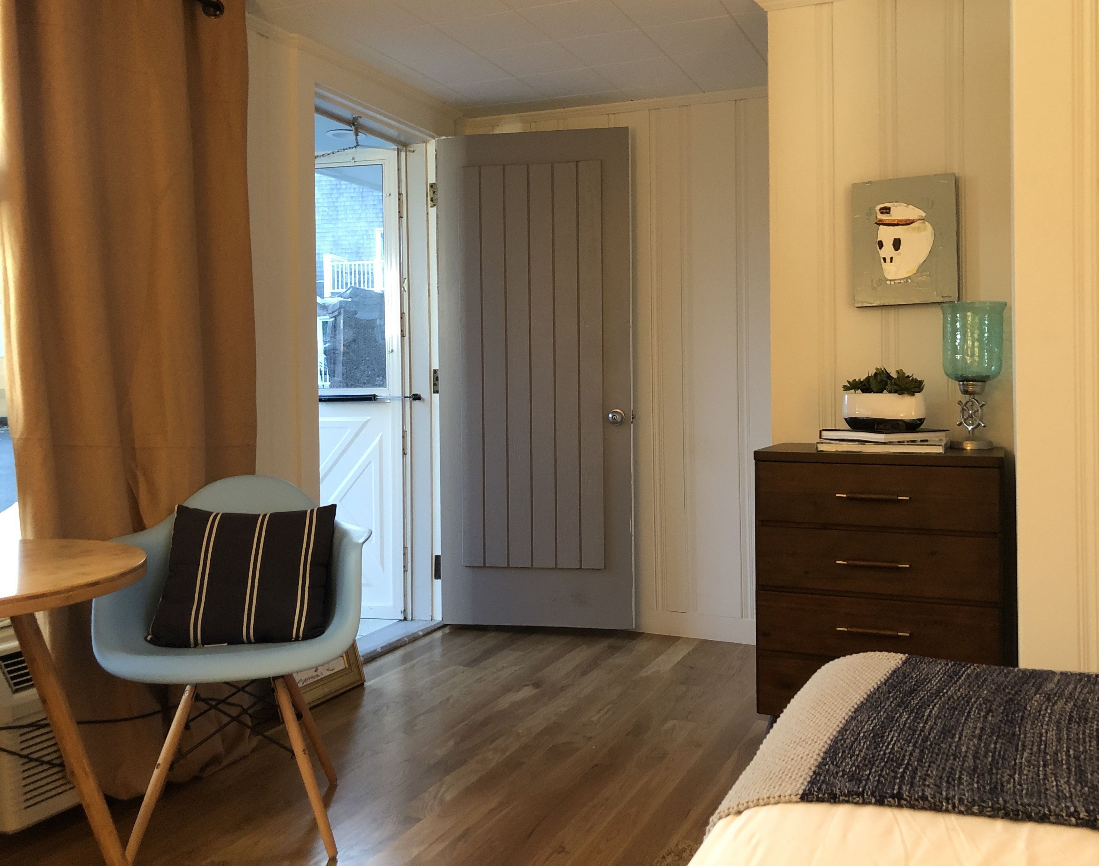 A room with a wooden floor, a blue chair with a striped pillow, a dresser with drawers, a small table, and a door leading to an outdoor area.