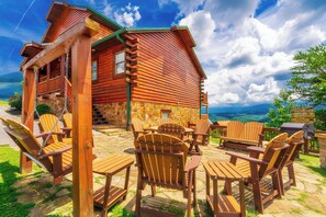 A wooden cabin with a stone foundation, outdoor seating area with wooden chairs and tables, and a scenic mountain view. - Majestic Mtn Lodge: Mtn Views, Theater, Games, Resort Pool, Hot Tub, Wifi (Sevierville)