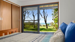 Ein Schlafzimmer mit einem großen Schiebeglastür, die zu einer grünen Fläche mit Bäumen und Blick auf das Meer führt.