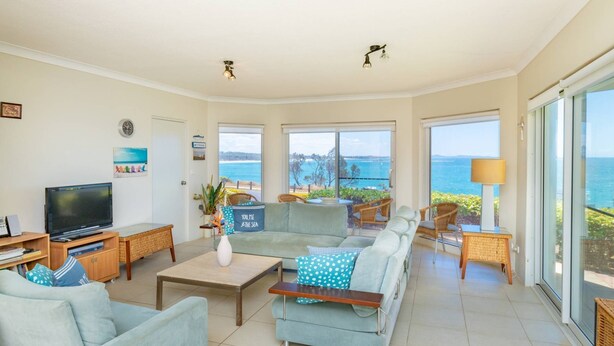A spacious living room with a large sofa, a flat-screen TV, and a view of the sea through large windows.