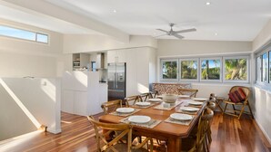 Un comedor moderno con una mesa de madera dispuesta para cuatro personas, rodeada de sillas de mimbre. Se ve una cocina con refrigerador y un comedor con vista a la vegetación a través de amplias ventanas.