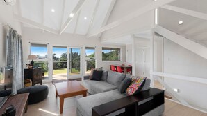 Amplia sala de estar con un sofá gris, una mesa de centro de madera y un mueble para televisión. Grandes ventanales ofrecen vistas al exterior.