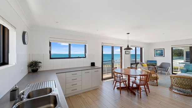 A modern kitchen with a large window, a dining area with wooden chairs, and a view of the ocean. - Jambi Ocean View - oceanfront Woolgoolga (Woolgoolga)