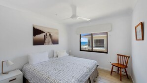 A bedroom with a bed, a chair, a ceiling fan, a window with a view, and a painting on the wall. - Jambi Ocean View - oceanfront Woolgoolga (Woolgoolga)