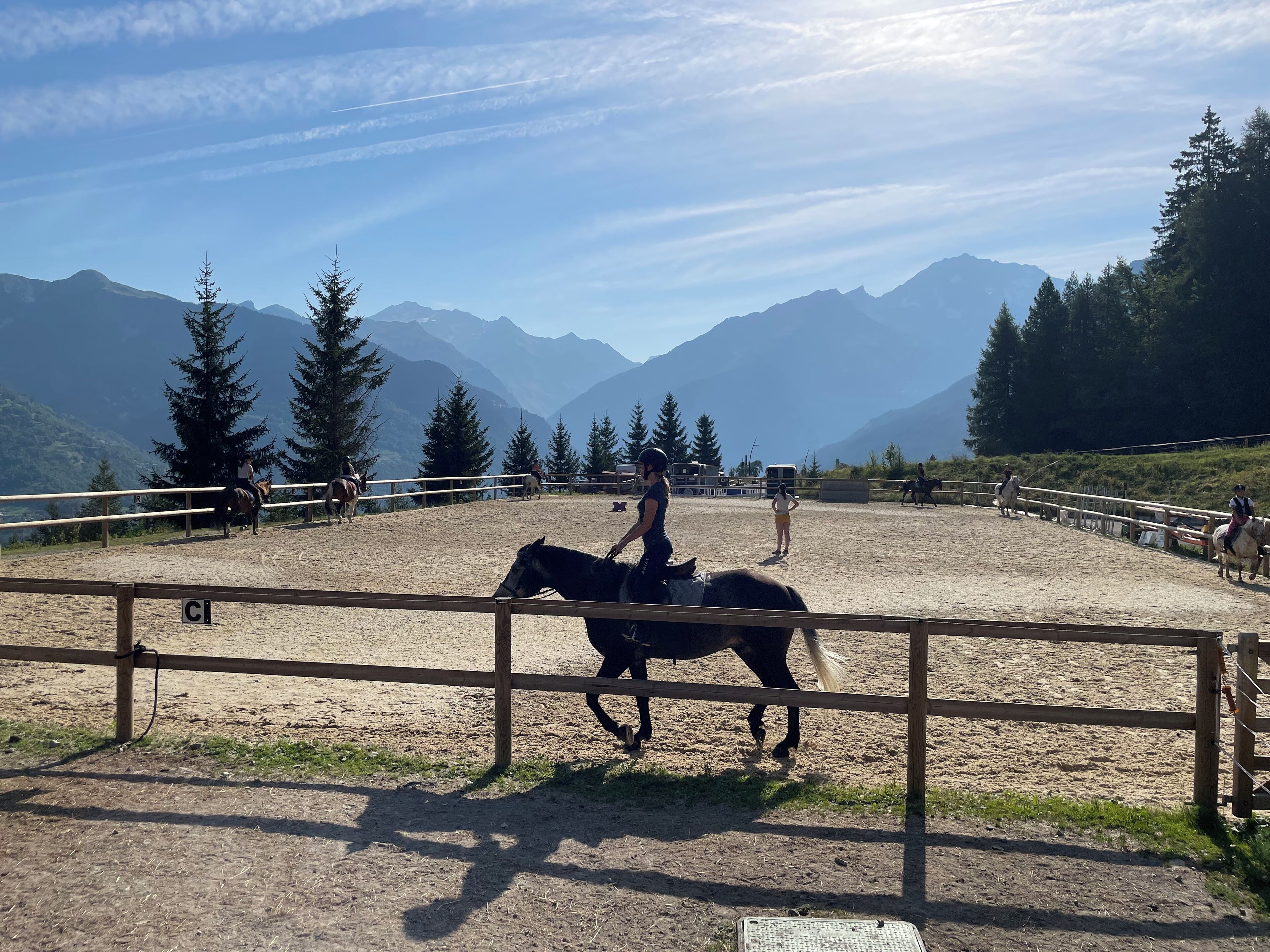 Un área ecuestre al aire libre con caballos y jinetes, rodeada de montañas y árboles.