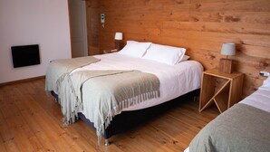A bedroom with a wooden wall, a bed with white linens, two bedside tables with lamps, and a flat-screen TV mounted on the wall. - Hotel Puerto Madera (Pelluhue)
