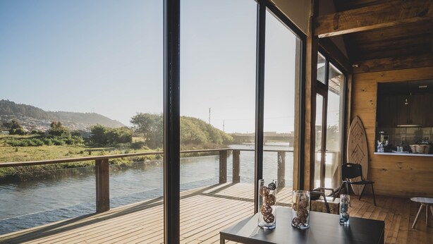 A wooden deck with a table and chairs overlooking a river, with a view of a distant town and hills. - Hotel Puerto Madera (Pelluhue)