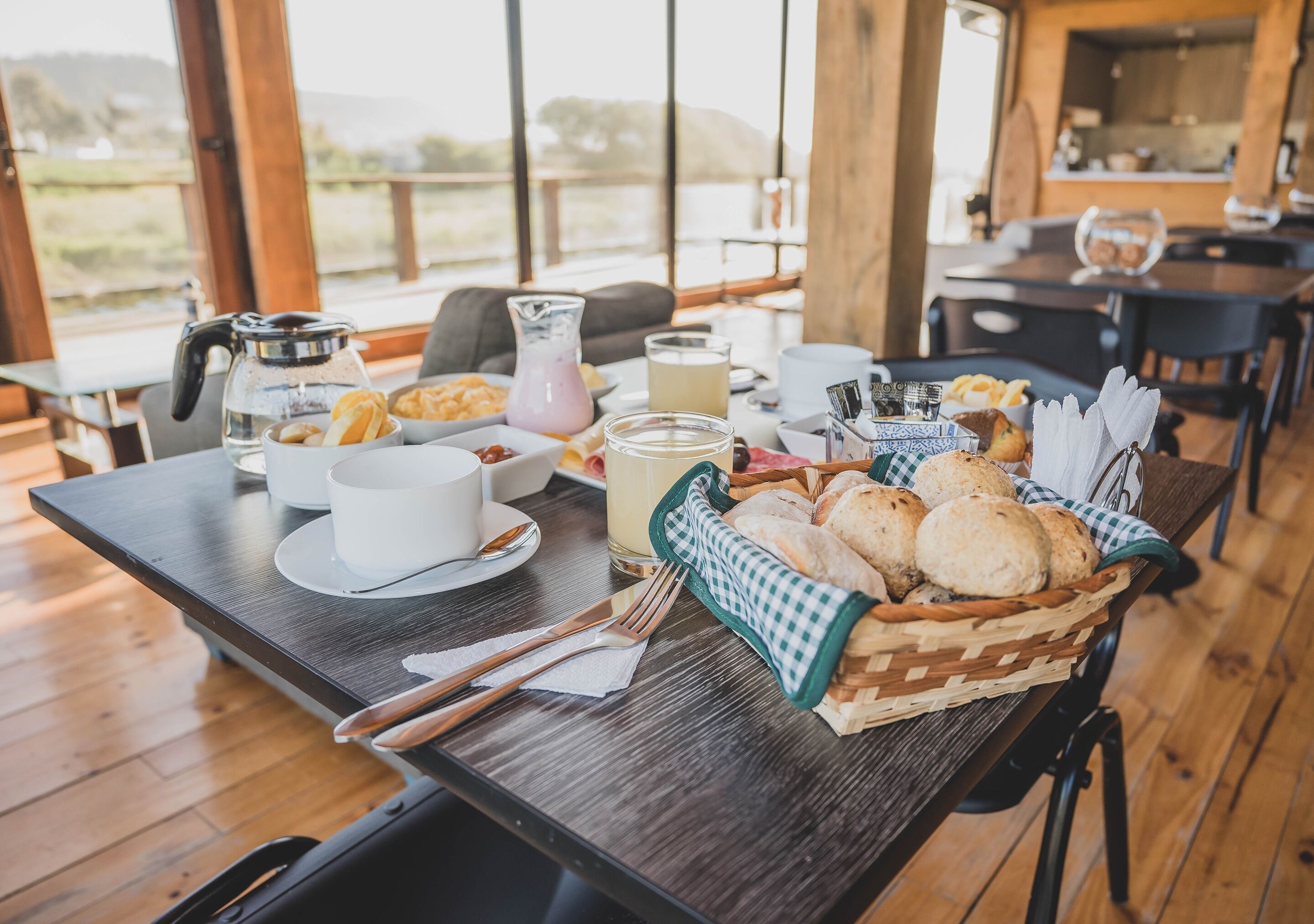 Una mesa de desayuno con pasteles, una jarrón y un vaso de leche, en una habitación con suelo de madera y ventanas amplias.
