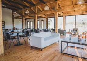 Un amplio salón con un sofá blanco, sillas negras y una mesa de comedor. La habitación cuenta con vigas de madera, ventanas grandes y vistas al exterior.