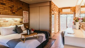Un dormitorio moderno con pared de ladrillo, una cama con ropa de cama blanca, una mesita de noche con una botella y flores, y un escritorio con una silla.