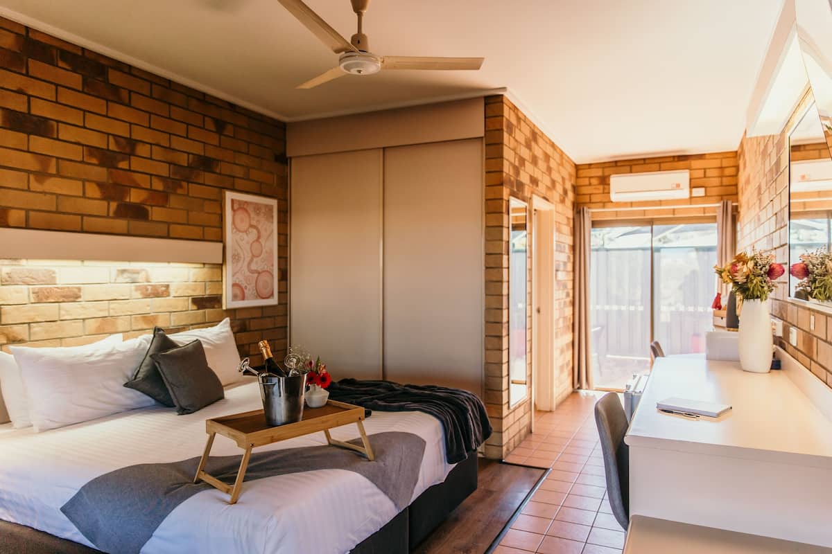 A modern bedroom with a brick wall, a bed with white bedding, a bedside table with a bottle and flowers, and a desk with a chair.