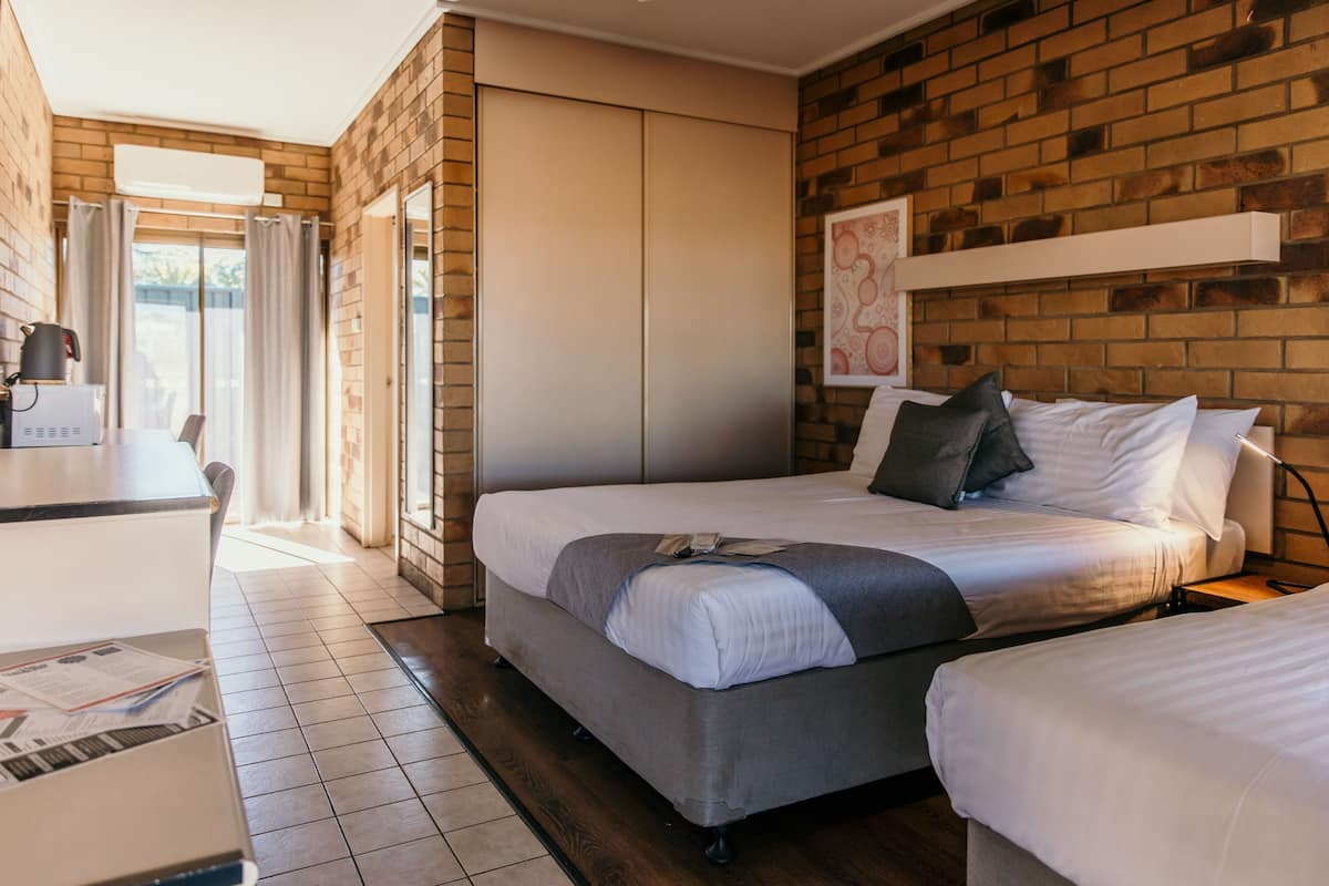 A modern hotel room with a brick wall, a large bed with white bedding, a desk with a kettle, and a view through a glass door.