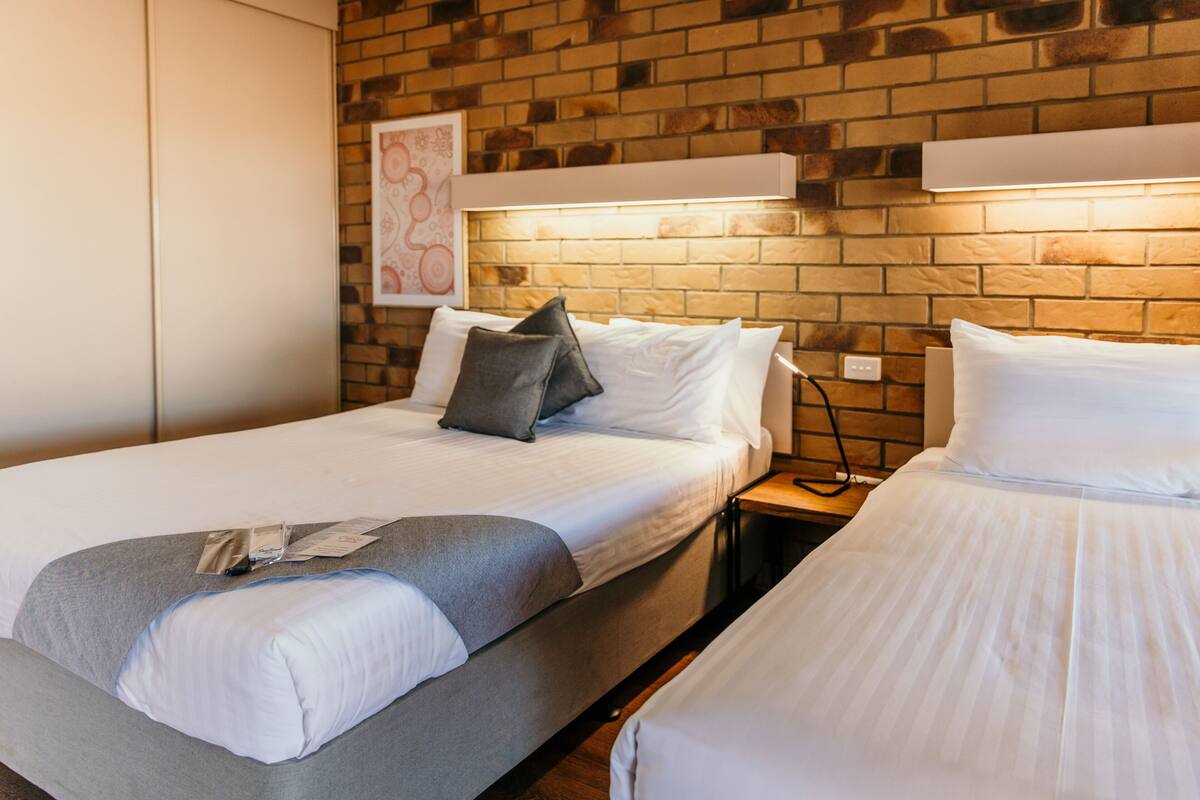 A hotel room with two beds, a brick wall, a framed artwork, and a bedside table with a lamp.