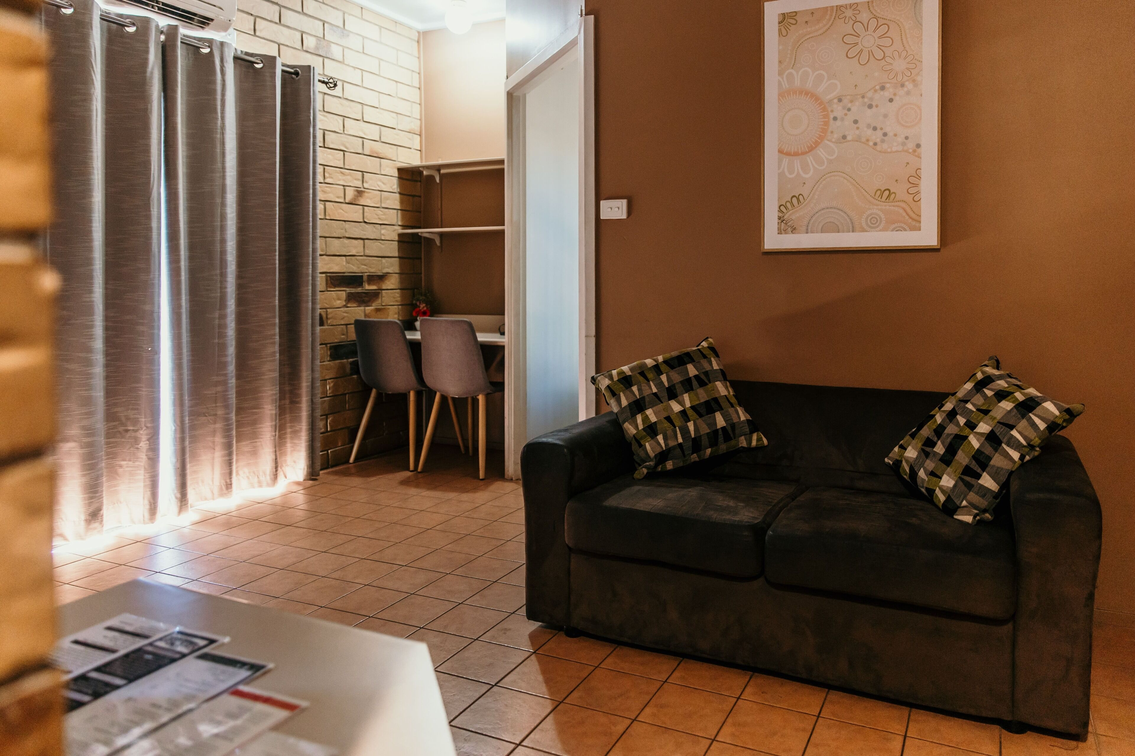 Una sala de estar con un sofá negro, dos cojines a cuadros, una mesa de comedor con sillas, una zona de cocina con pared de ladrillo y una obra de arte grande enmarcada en la pared.