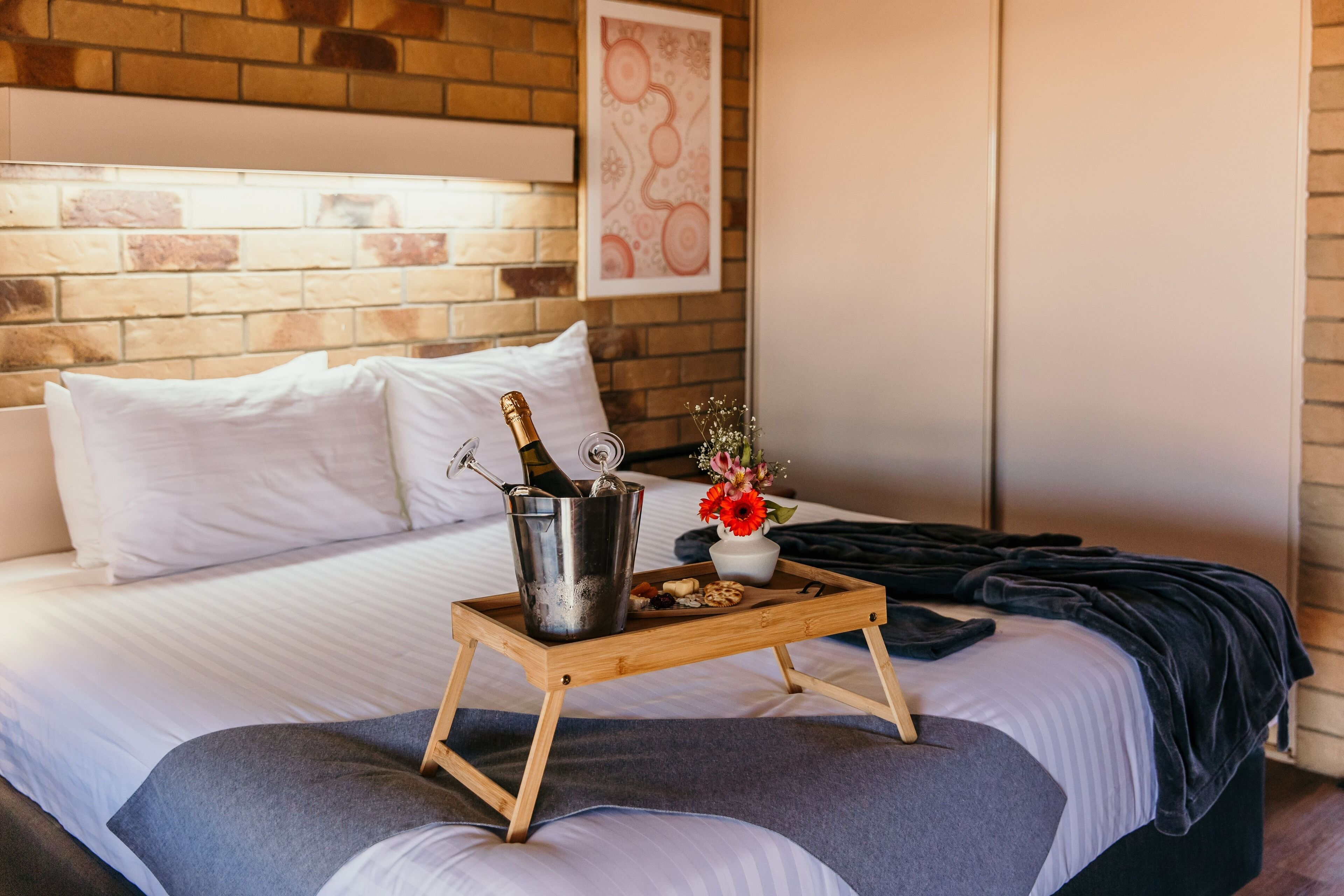 Una cama bien hecha con una bandeja de madera que sostiene una botella, un vaso y un pequeño arreglo floral.