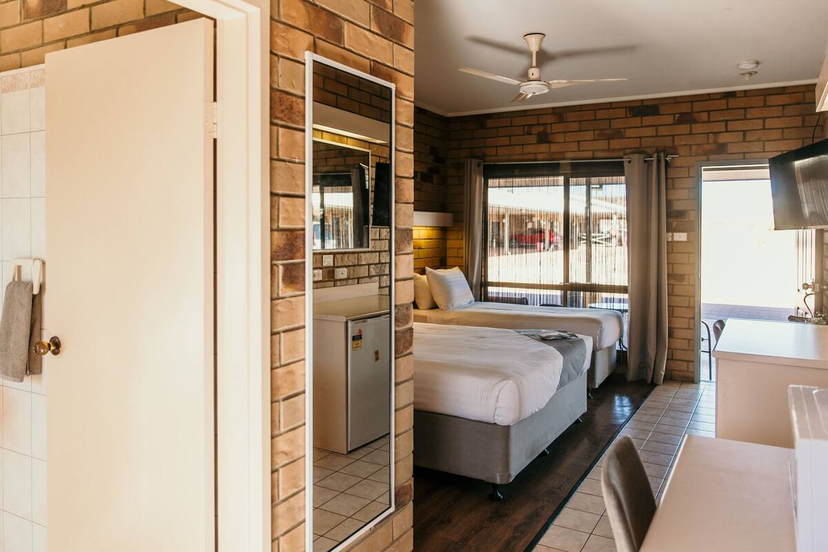 A hotel room with a brick wall, two beds, a desk, a small refrigerator, and a ceiling fan.