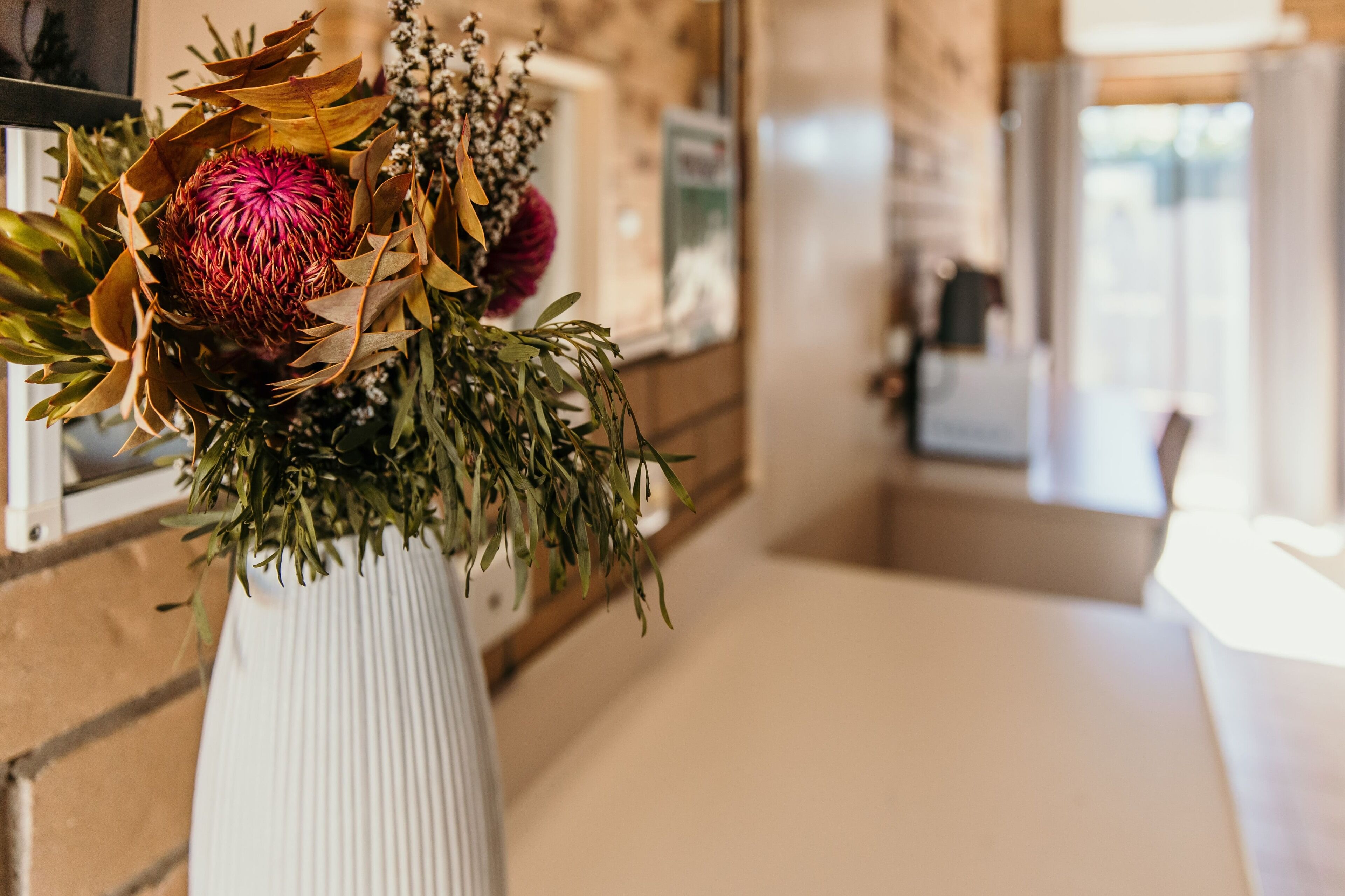 Un vase blanc orné d’une composition florale comprenant une fleur rouge bien visible, placé sur un comptoir dans un couloir bien éclairé, avec un mur en bois et une porte en arrière-plan.