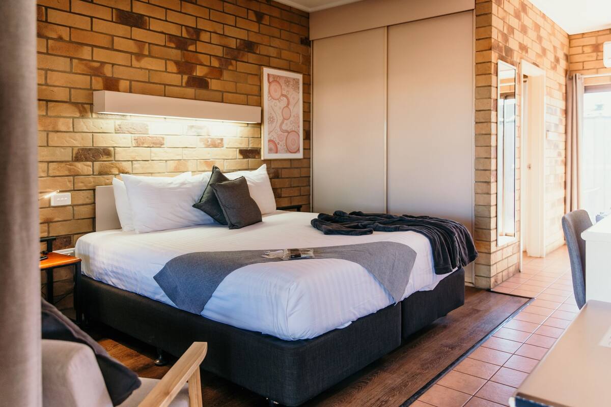 A modern hotel room with a brick wall, a large bed with white and grey bedding, a bedside table, and a chair.
