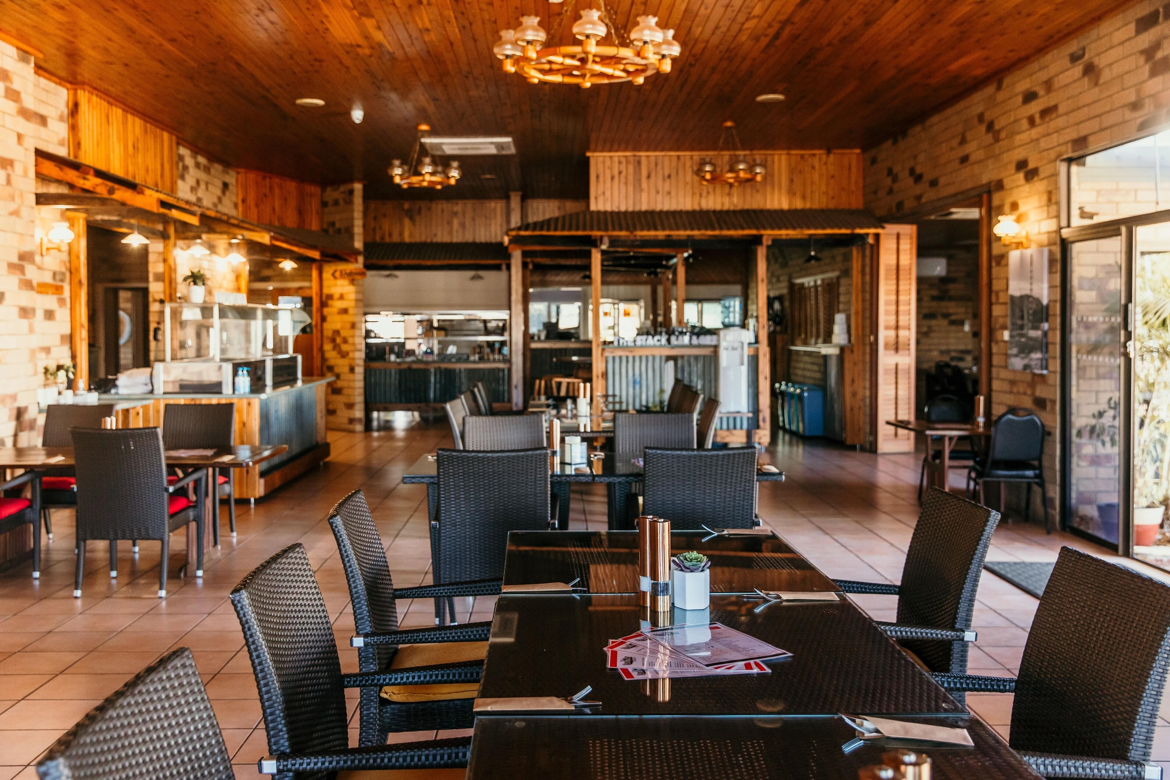 A restaurant with a rustic interior, featuring brick walls, wooden ceilings, and a mix of black and wicker furniture.