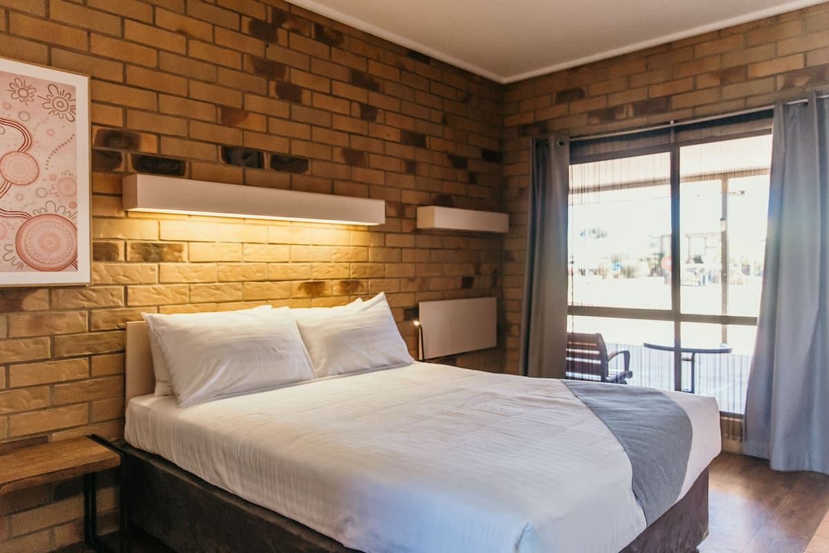A modern hotel room with a brick wall, a large bed with white bedding, a bedside table, a wall-mounted light, and a view through a large window.