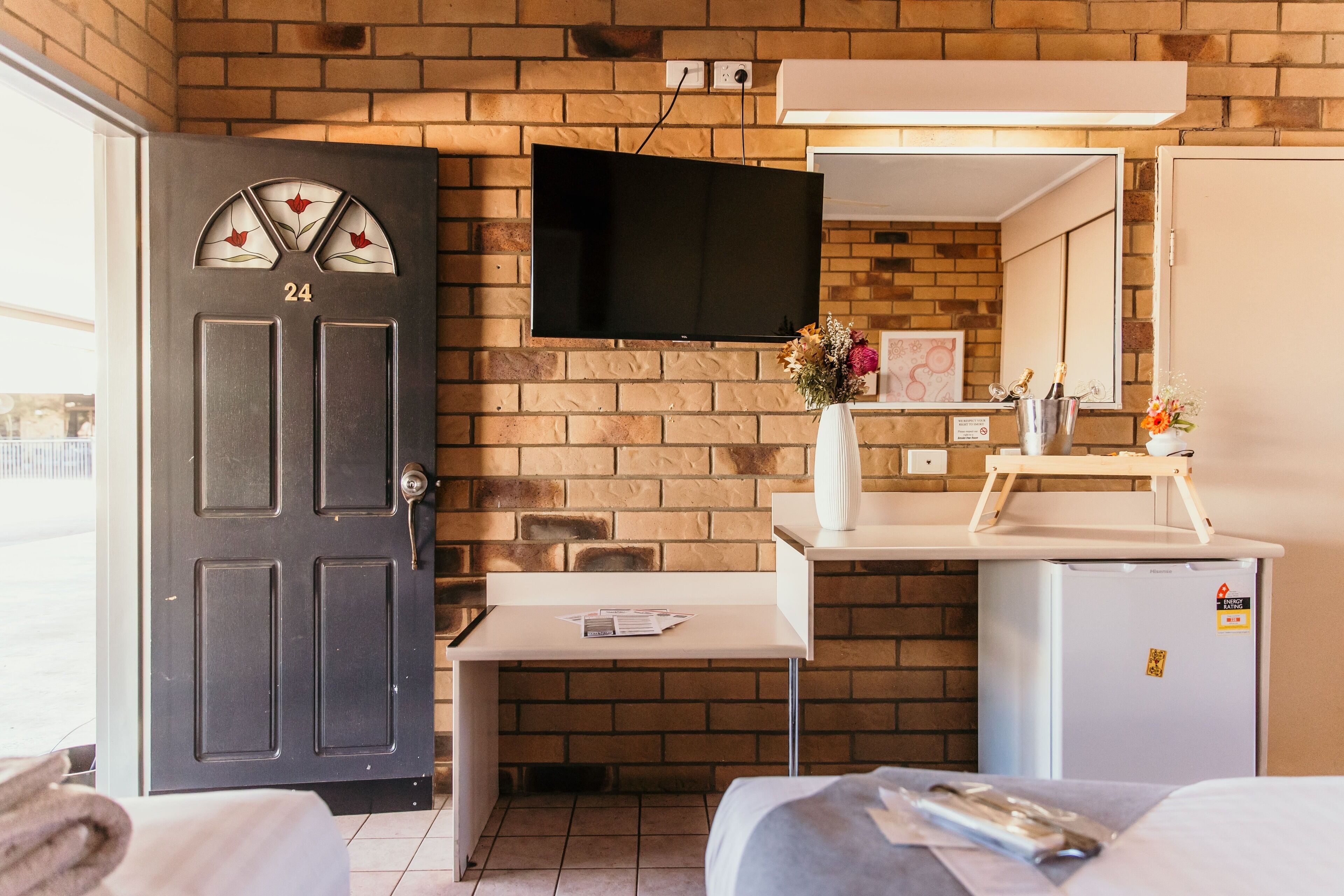 A compact hotel room with a brick wall, a flat-screen TV mounted above a desk, a small refrigerator, and a door with a number plate.
