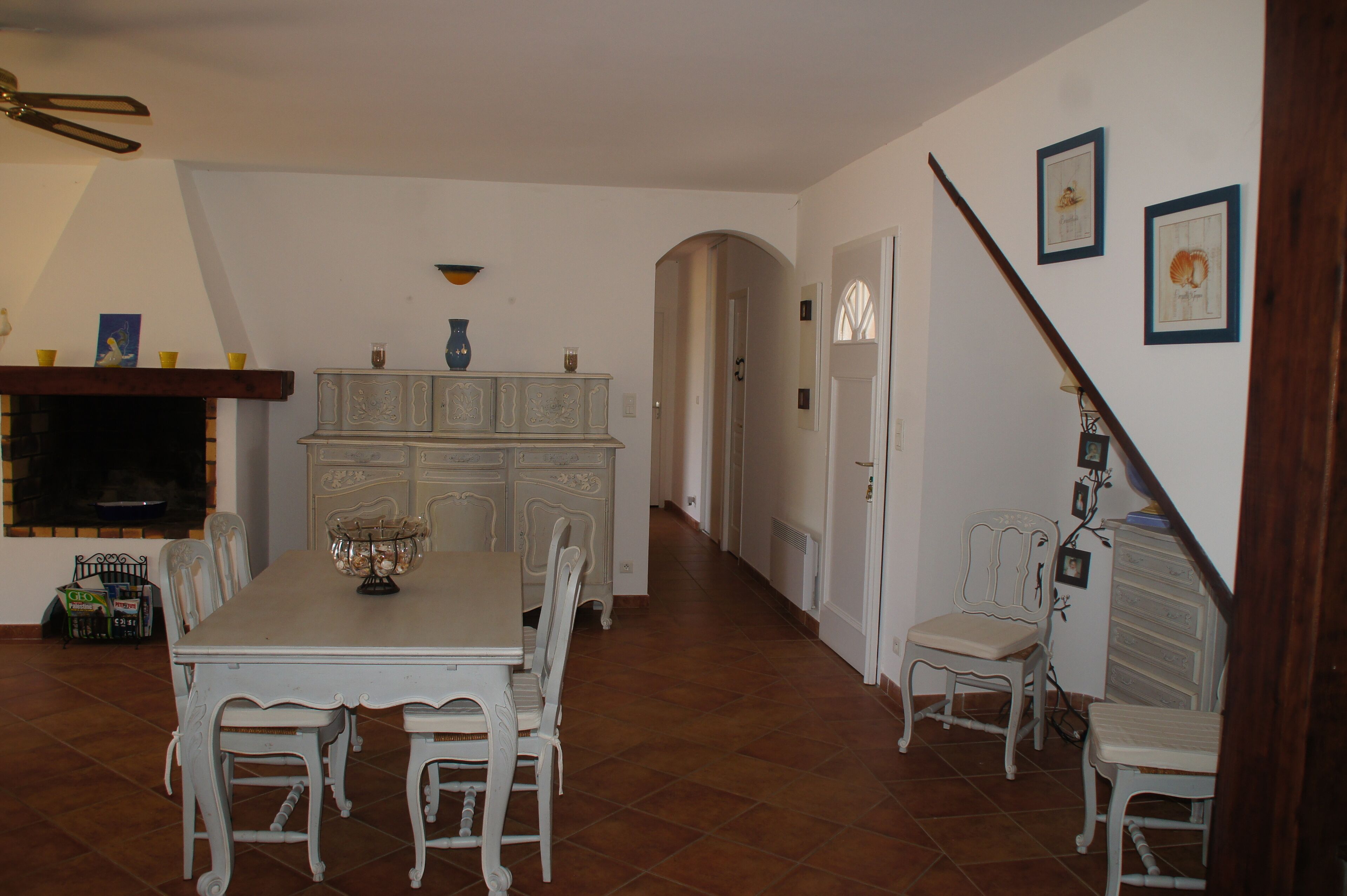 Un coin repas avec une table et des chaises blanches, une cheminée et un escalier menant à une autre pièce.