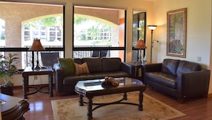 A living room with a sofa, coffee table, and a ceiling fan. There is a window with a view of palm trees and a building. - Great Getaway at Shores at Waikoloa! (Waikoloa)