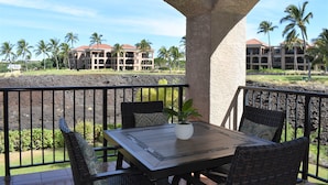 A covered outdoor seating area with a table and chairs, overlooking a pool and a building with a red roof. - Great Getaway at Shores at Waikoloa! (Waikoloa)