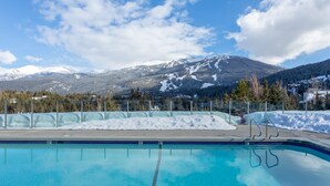 A swimming pool with clear blue water, surrounded by a metal fence, with a snowy mountain and forested area in the background. - SUPER SUMMER GETAWAY  Views + POOL + HOT TUB (WD, FP) (Whistler)