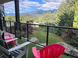 A balcony with a view of mountains, a red chair, and a wooden table with magazines. - SUPER SUMMER GETAWAY  Views + POOL + HOT TUB (WD, FP) (Whistler)