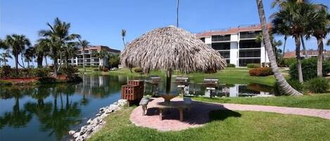 A resort with a thatched-roof gazebo, a pond, palm trees, and a building with a red roof in the background.