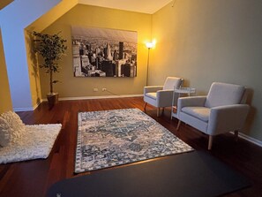 Una sala de estar con un cuadro de un paisaje urbano, dos sillones blancos, un tapete estampado, una planta en maceta y una lámpara de pie.