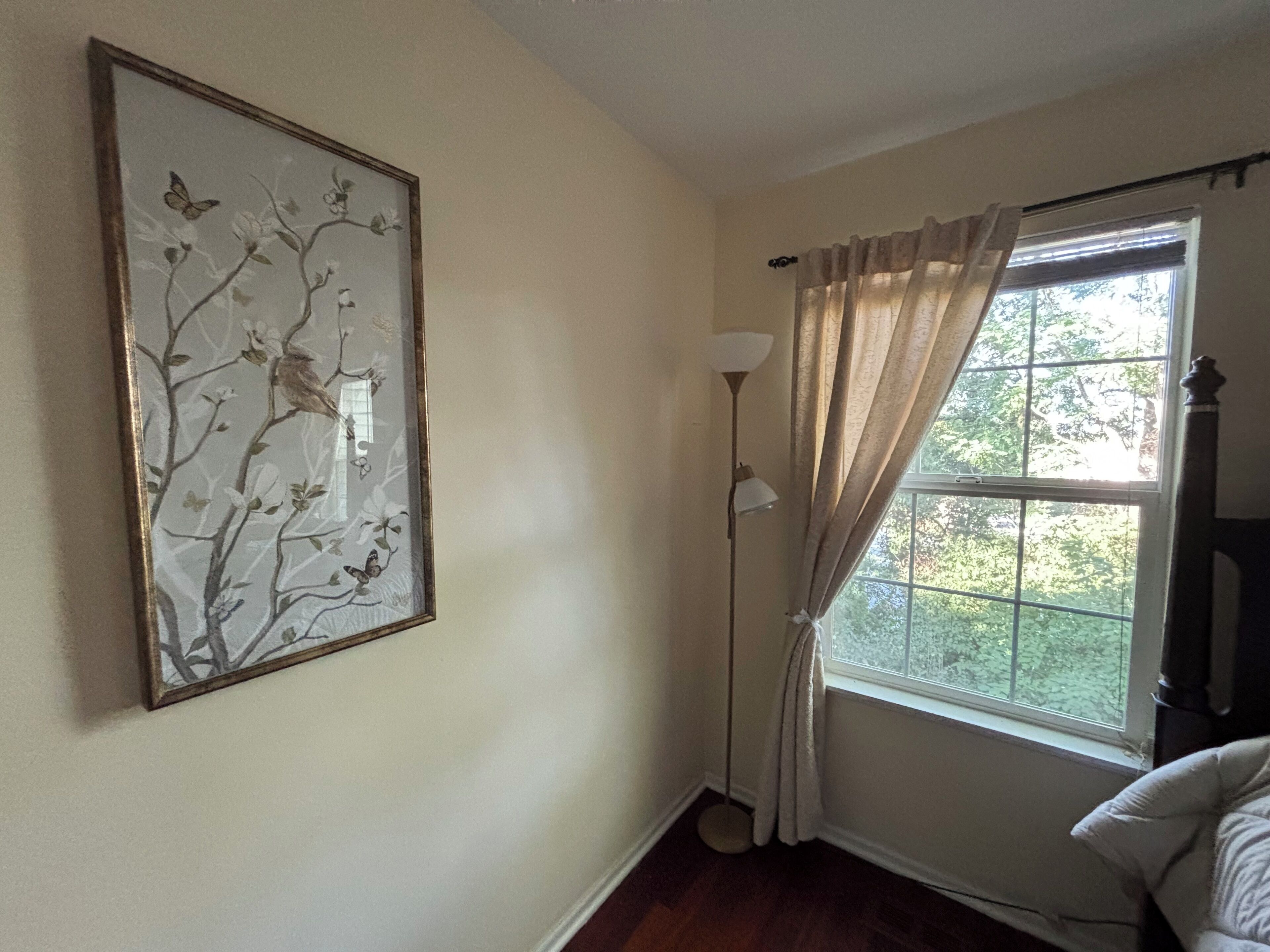 Una stanza con un quadro floreale appeso al muro, una lampada da terra, una finestra con tende e un letto.