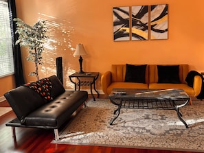 A living room with a black leather sofa, a patterned rug, a coffee table, a side table with a lamp, and abstract wall art.