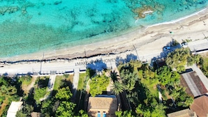 An aerial view of a beach with clear turquoise waters, a sandy shore, and a row of houses with a small building in the foreground. - Makis&Fani Homes (Lefkada)
